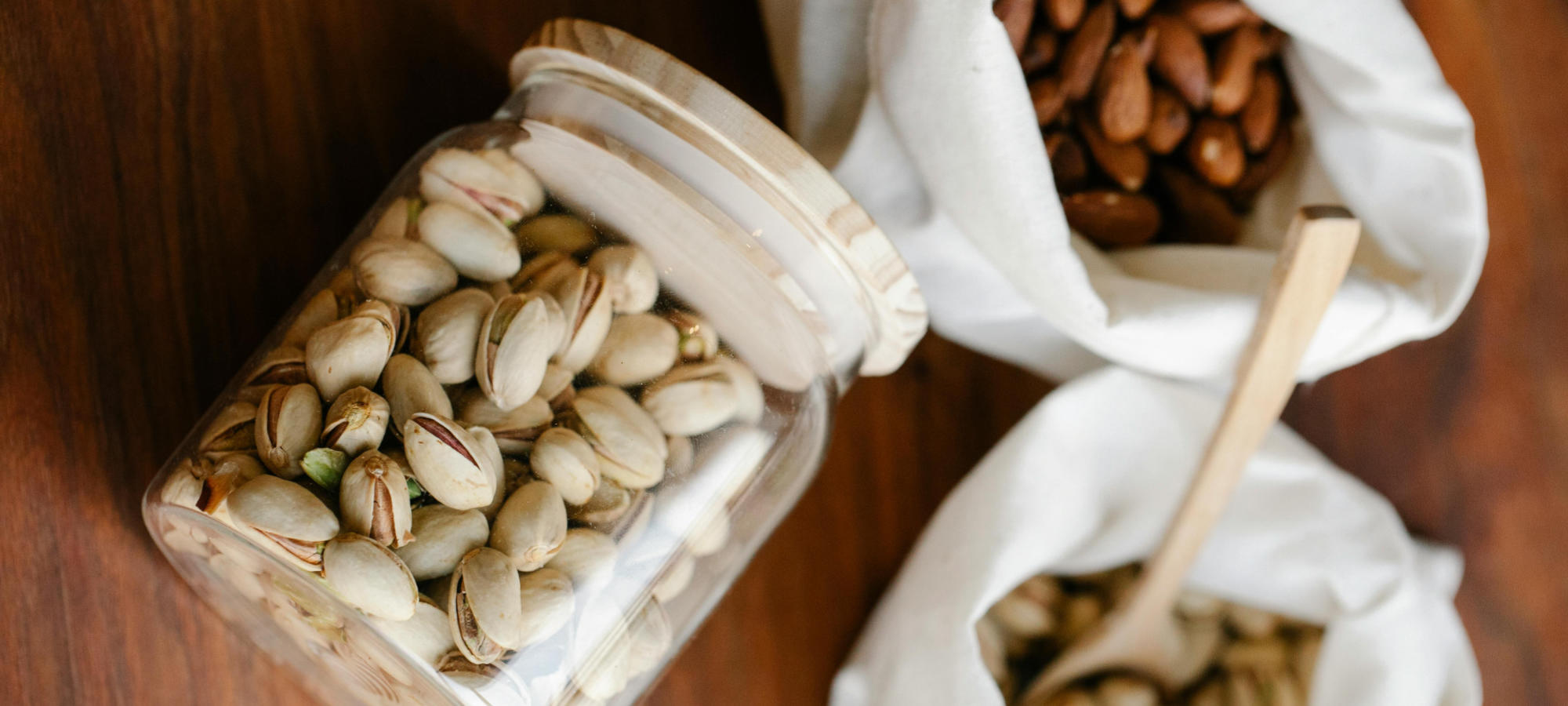 pistacchi in guscio dentro un barattolo con tappo ermetico in legno, altri pistacchi in guscio dentro un sacchetto di tela con cucchiaio in legno e un altro sacchetto di tela pieno di mandorle sgusciate